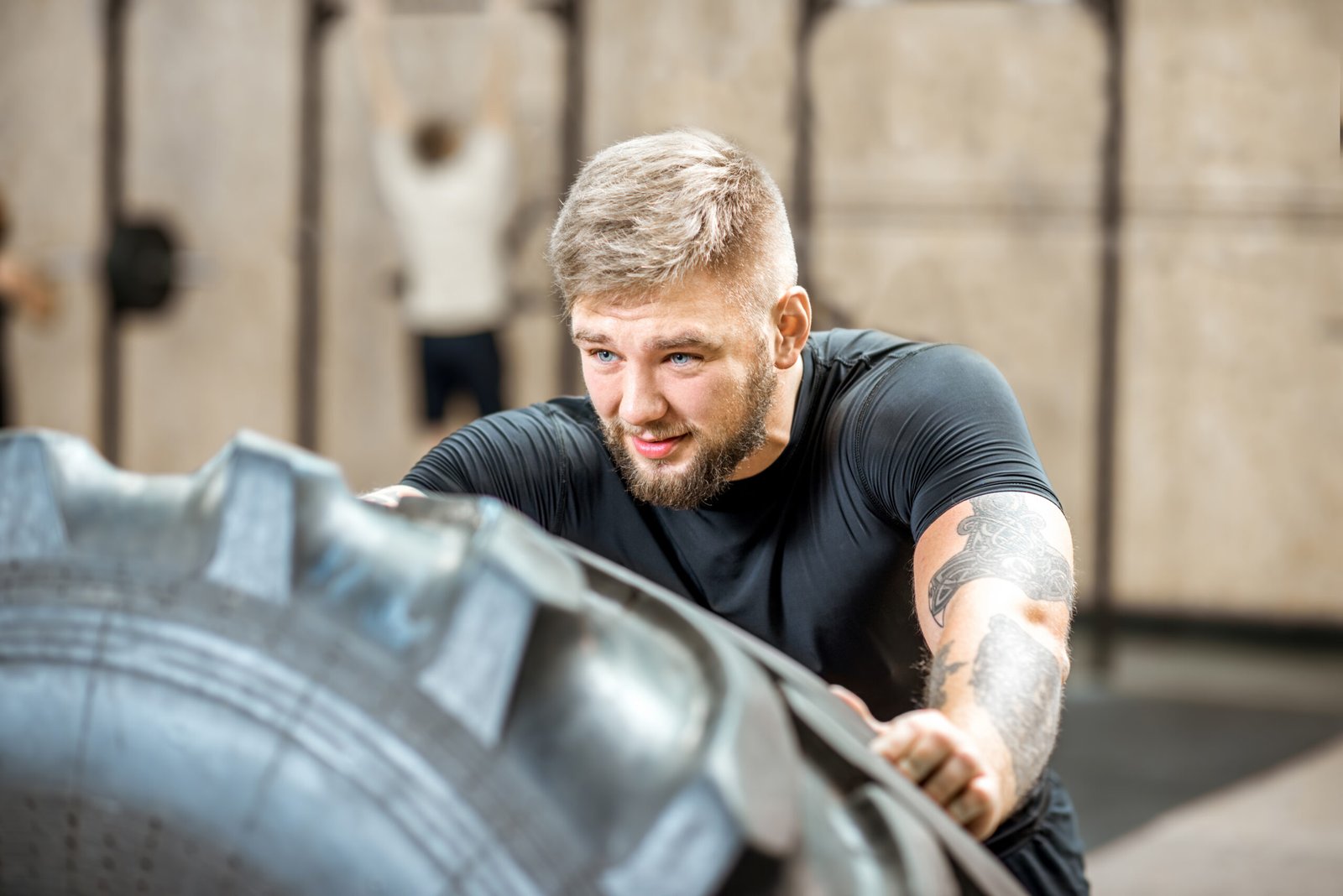 Athletic man in black sportswear pushing a big tire training in the gym